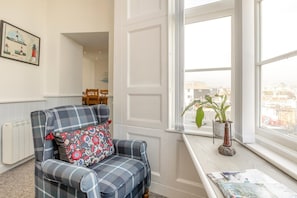 Ein gemütliches Zimmer mit einem Karo-Sessel, einer Heizung, einem Fenster mit Blick auf die Stadt sowie einem Tisch mit Pflanze und Zeitschrift.
