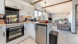 A modern kitchen with white cabinets, a built-in oven, and a coffee machine. There is a dining area with a gray sofa and a small table. - 2 Bed in Llangain (oc-g30185) (Llangain)
