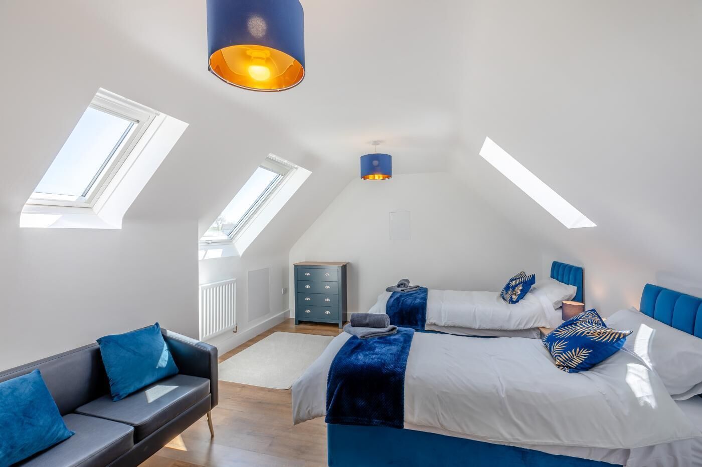 A bedroom with a sloped ceiling, skylights, a bed with blue bedding, a gray sofa, and a dresser.