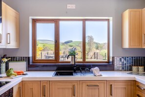 A kitchen with wooden cabinets, a large window with a view of rolling hills, a sink, and a countertop with kitchen utensils. - 5 Bed in Pant-y-Dwr (oc-w30200) (Pant-y-dwr)