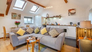 Een moderne woonkamer met een grijze bank, een klein bijzettafeltje en een keuken met witte kasten en een houten aanrecht.
