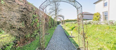 A pathway lined with a metal archway, flanked by a hedge and a building.