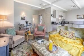 Una sala de estar con un sofá, un sillón y una mesa de centro con un libro y una jarrón. Hay una zona de comedor con una mesa y sillas, y una cocina al fondo.