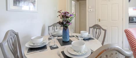 A dining room with a round table set for four, surrounded by chairs, a vase with flowers, and a framed picture on the wall.