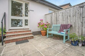 Un patio con un banco de madera, una planta en maceta con flores rosadas y una escalera de ladrillo que lleva a una puerta.