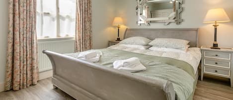 A neatly made bed with a green blanket and two rolled towels, a bedside table with a lamp, and a mirror on the wall.