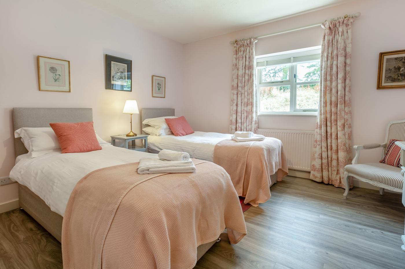 A hotel room with two beds, a nightstand with a lamp, a window with curtains, and a chair.
