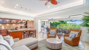 A spacious living area with a sofa, two wicker chairs, a coffee table, and a kitchen with a central island. - Luxurious Ho'olei 12-2, Lush View, Close to Pool, Sleeps 8 in Beds, Central AC! (Kihei)
