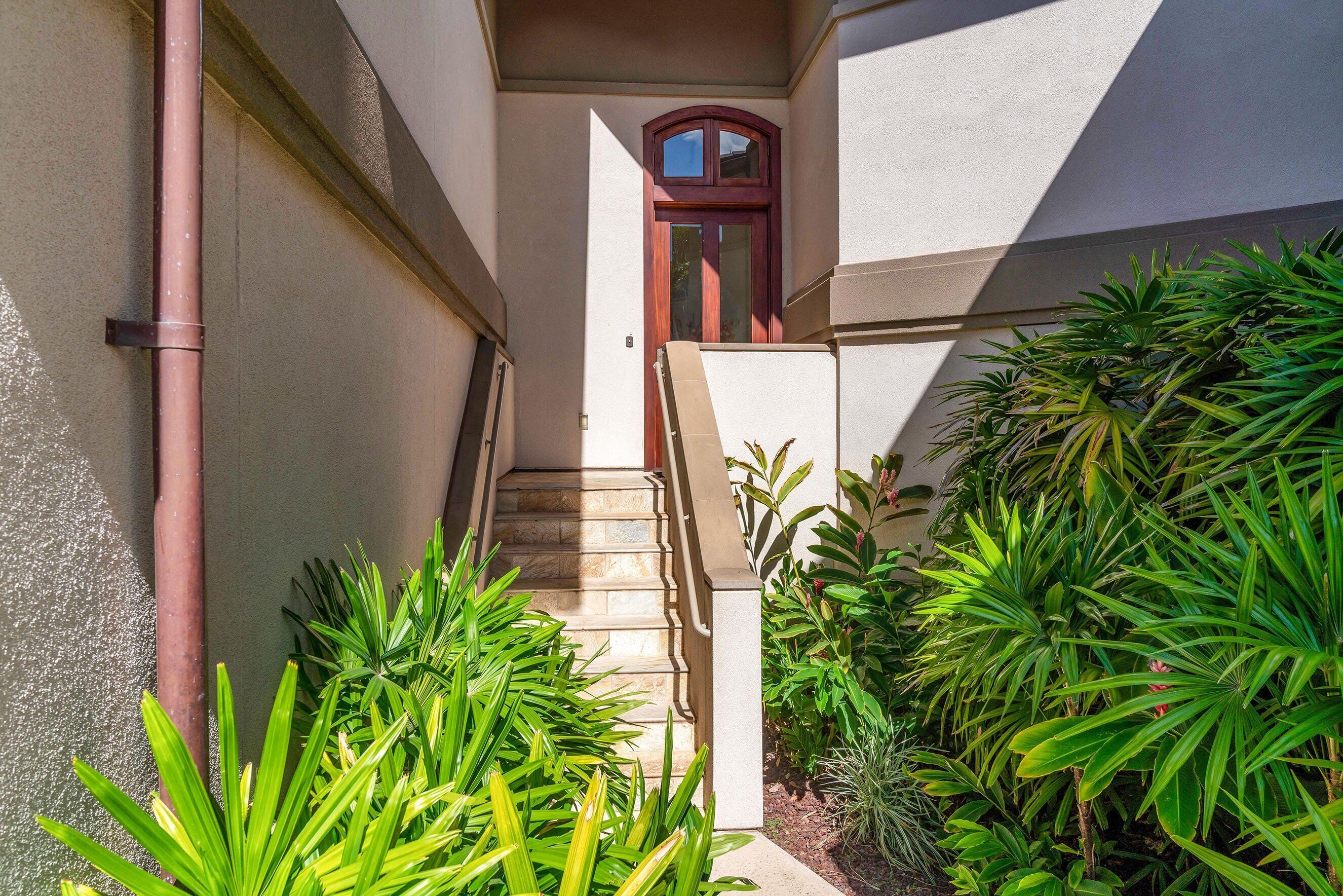 A narrow outdoor corridor with a staircase leading to a door, surrounded by lush green plants.