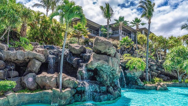 A resort with a natural rock waterfall, clear blue pool, and tropical vegetation. - Luxurious Ho'olei 12-2, Lush View, Close to Pool, Sleeps 8 in Beds, Central AC! (Kihei)