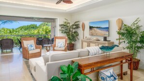 A spacious living area with a white sofa, wicker chairs, a wooden coffee table, and a view of the ocean through a sliding door. - Luxurious Ho'olei 12-2, Lush View, Close to Pool, Sleeps 8 in Beds, Central AC! (Kihei)