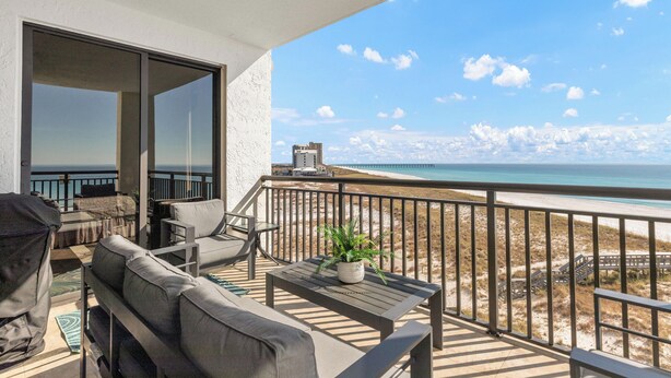 Ein Balkon mit einer Couch, Stühlen, einem kleinen Tisch und einem Blumentopf. Der Balkon bietet Blick auf einen Strand mit einem Pier im Hintergrund.