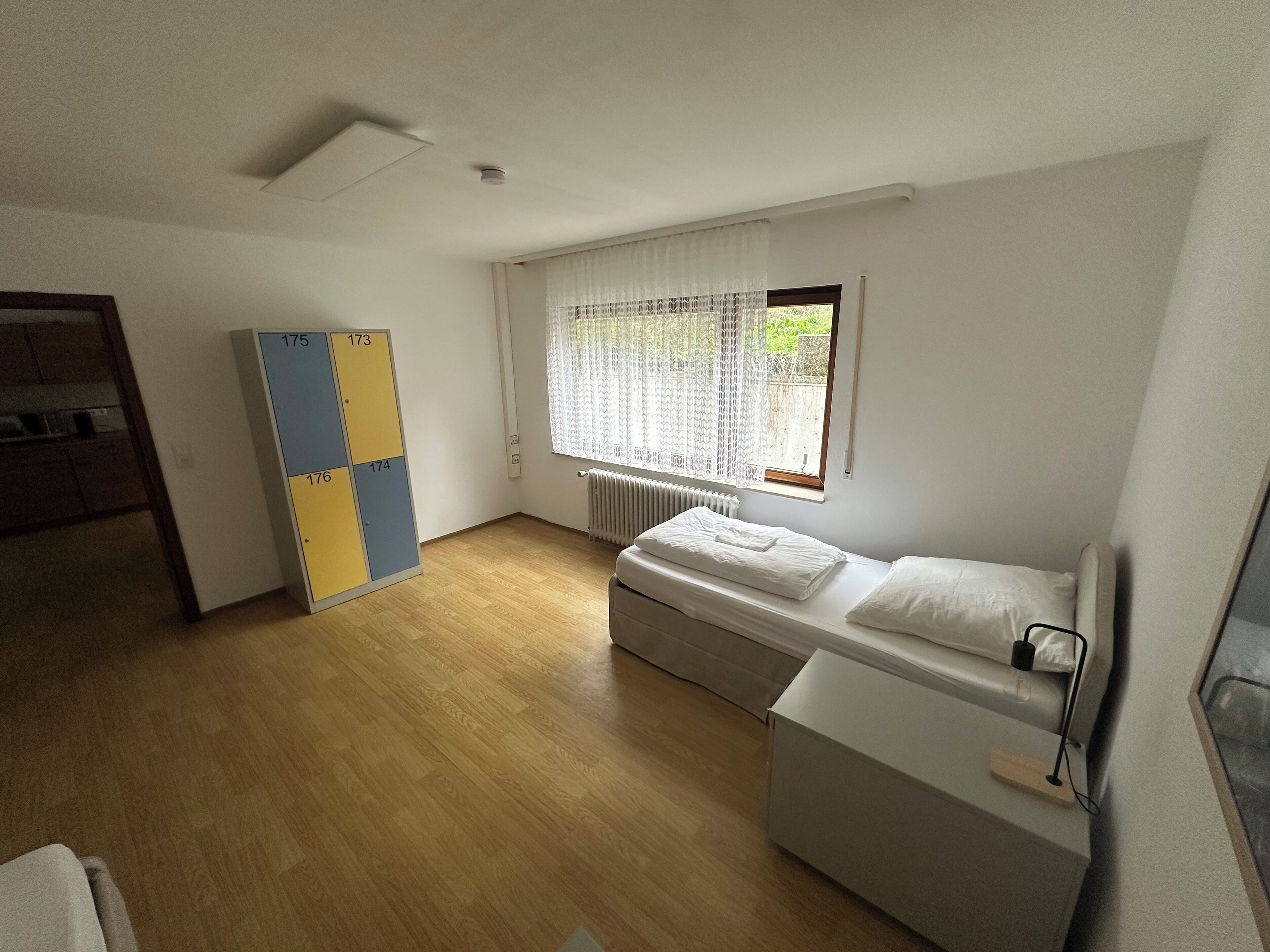 Quarto pequeno e limpo com cama, radiador e janela com cortinas.