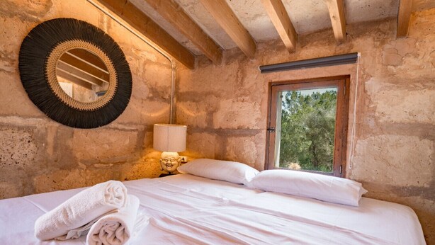Un letto con lenzuola bianche, uno specchio rotondo, una lampada e una finestra vista alberi.