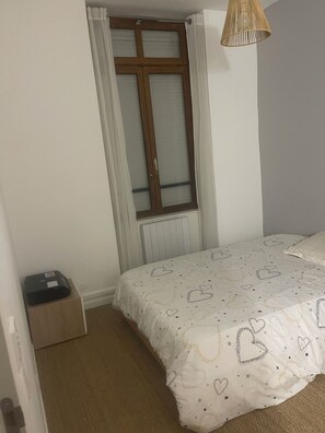 A bedroom with a bed, a window with curtains, a radiator, and a small box on the floor. - Room for rent in a private home (Caluire-et-Cuire)