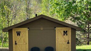Pequeña construcción de madera con dos puertas señalizadas con 'W' y 'M', dos sillas en el frente, y un camino de grava que la conduce.