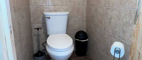A small bathroom with a toilet, a black trash bin, and a metal rack holding toilet paper.
