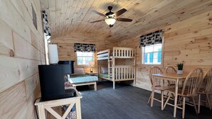 Interior de una cabaña con una cama litera, un escritorio con computadora, una mesa de comedor con sillas y un ventilador de techo.