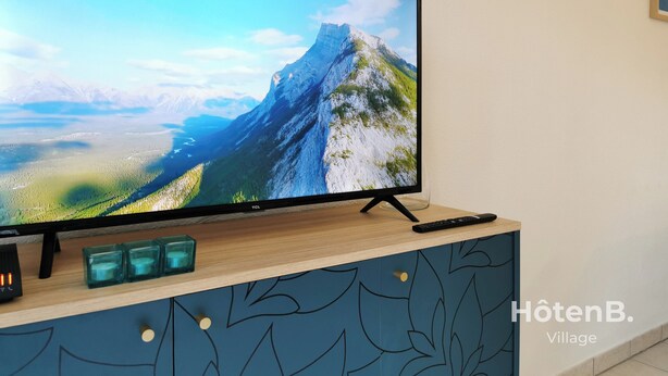 A flat-screen TV on a wooden stand with a mountain view, a remote, and three teal candle holders. - “Balcons de Renoir” Residence – Parking & Pool (Limoges)