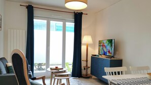 A room with a sofa, a dining table, a TV, and a lamp. - “Balcons de Renoir” Residence – Parking & Pool (Limoges)