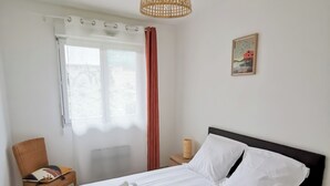 A neatly made bed with white linens and two rolled towels, a chair with a book, a small table with a candle, and a window with a view of greenery. - “Balcons de Renoir” Residence – Parking & Pool (Limoges)