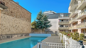 A swimming pool with a stone wall on one side and a modern building on the other. - “Balcons de Renoir” Residence – Parking & Pool (Limoges)