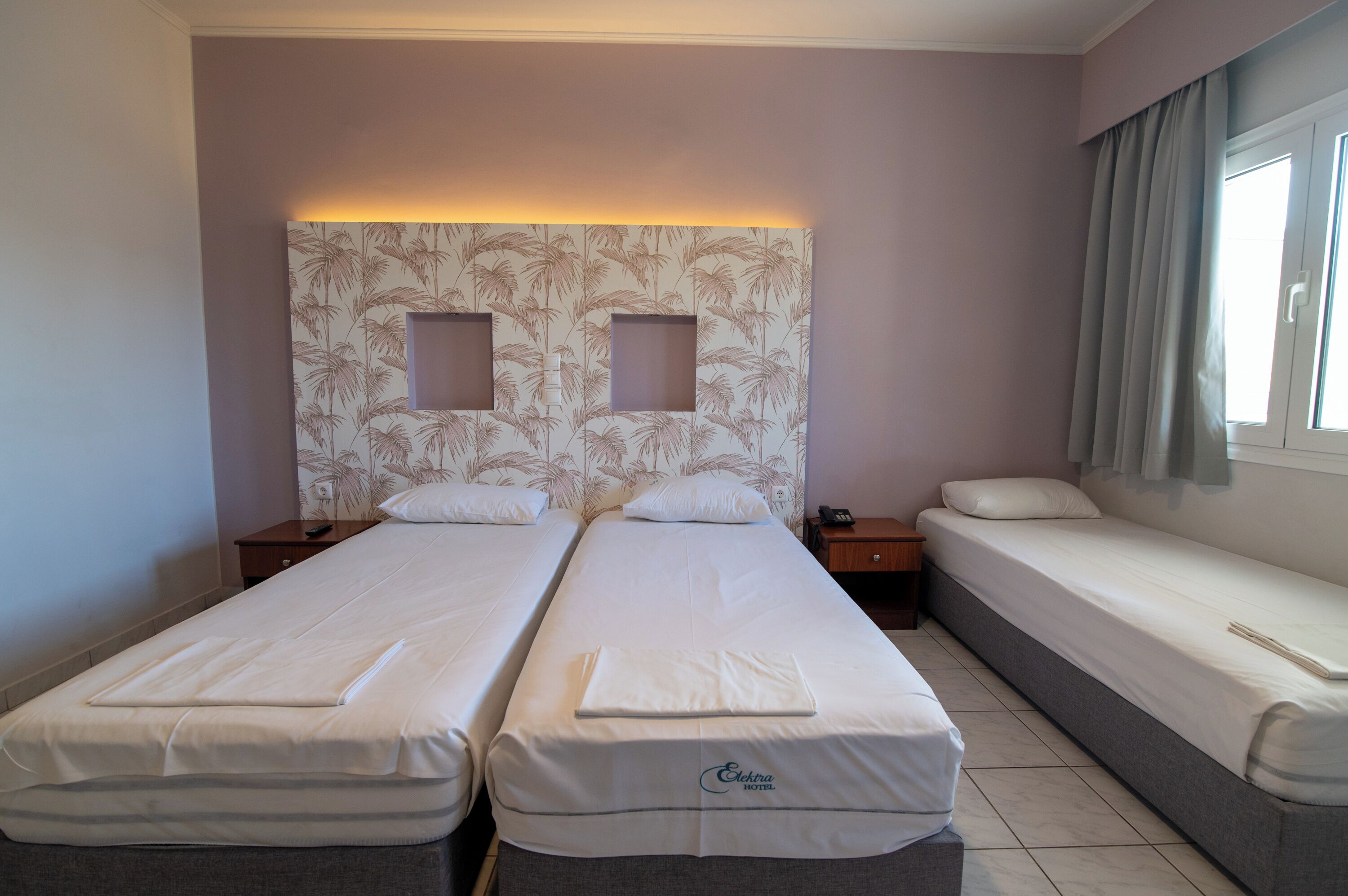 A hotel room with two single beds, a headboard with a leaf pattern, a nightstand with a phone, and a window with curtains.