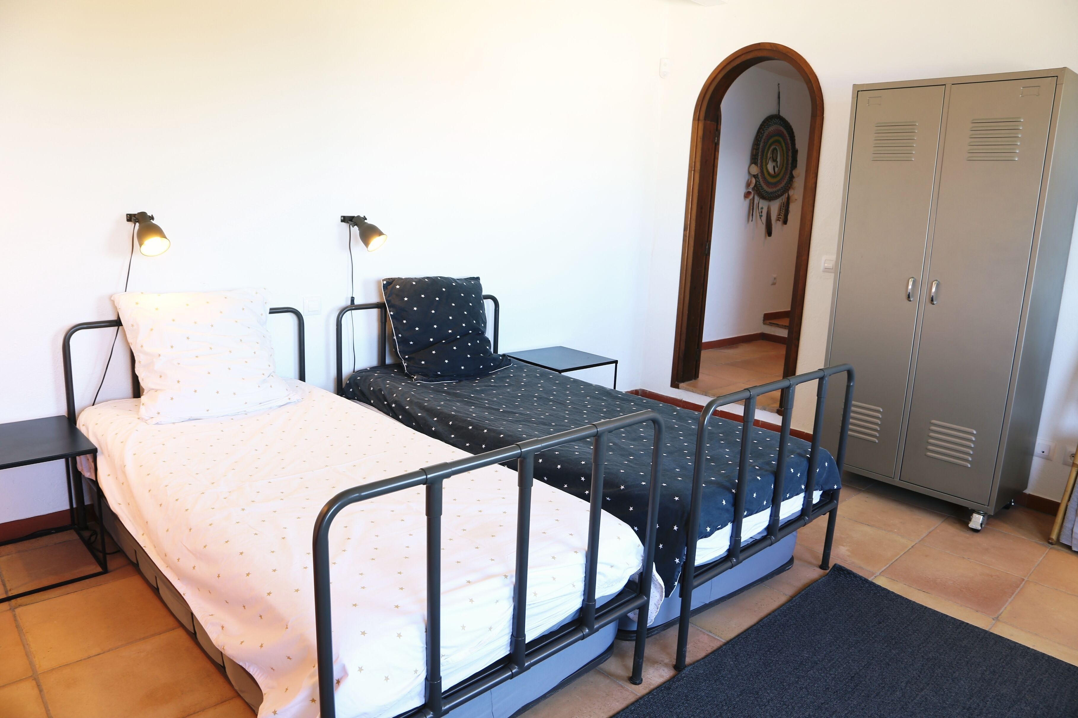 A single bed with a metal frame, a bedside table, a lamp, a wardrobe, and a mirror.