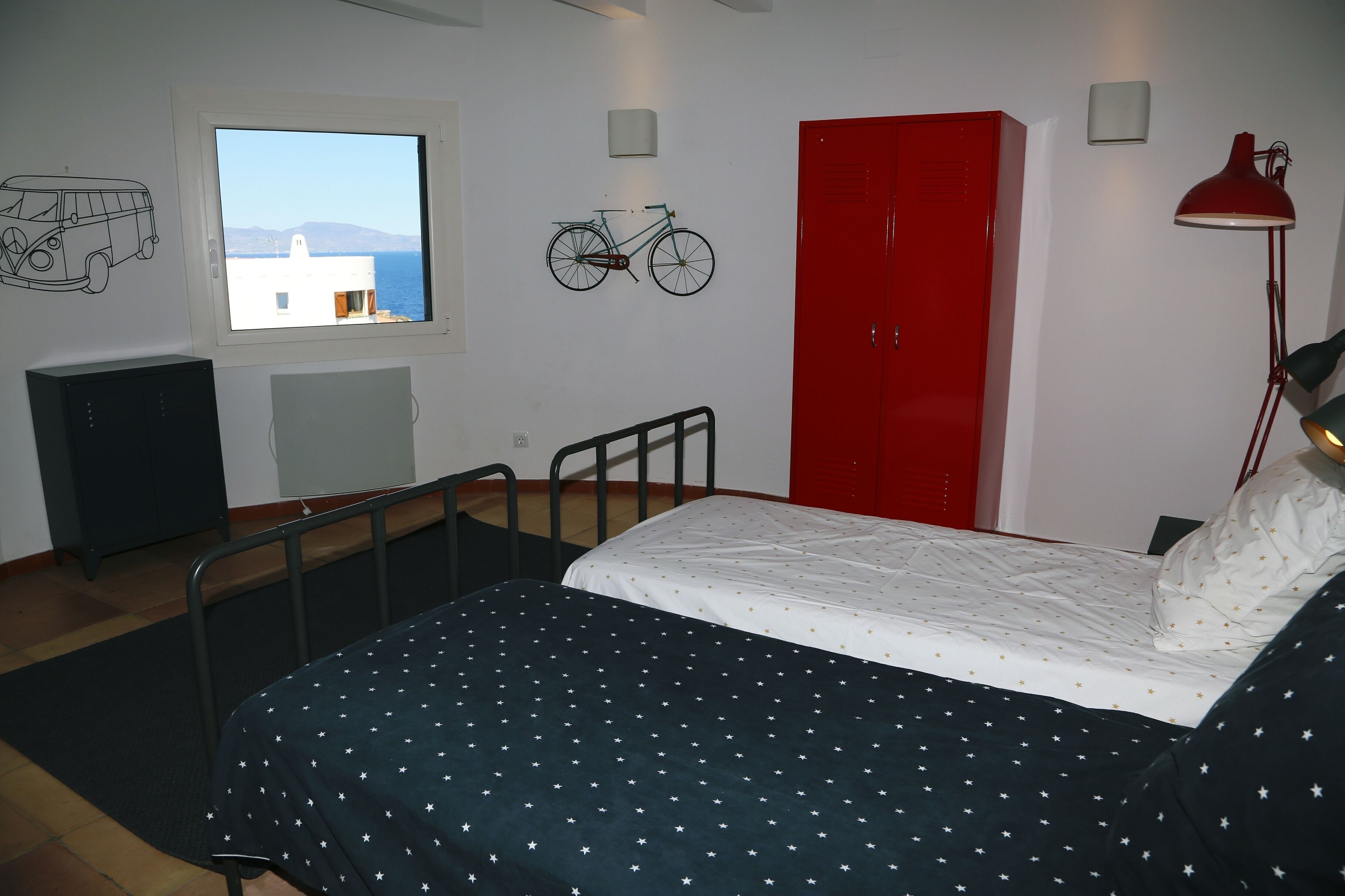 A bedroom with a bed, a window with a view, a red wardrobe, a bicycle wall art, and a desk.