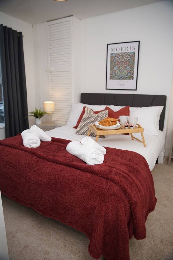 Un lit bien fait, recouvert d’une couverture rouge, un plateau avec des articles pour le petit-déjeuner et un tableau encadré accroché au mur.