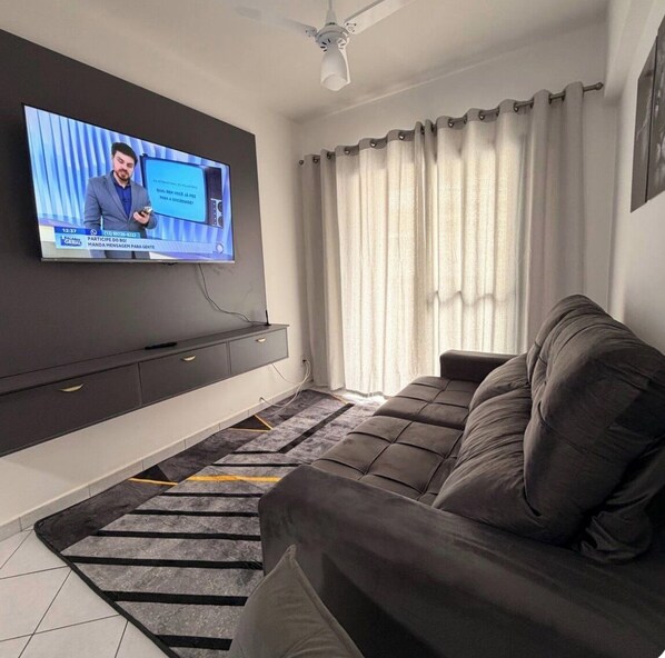 A modern living room with a large flat-screen TV mounted on the wall, a dark sofa, and a staircase with a patterned rug. - Apartment facing the beach in Aviação, Praia Grande (Praia Grande)