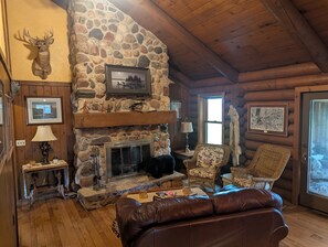 Ein Blockhaus-Interieur mit Kamin aus Naturstein, Holzwänden, einem aufgehängten Hirschgeweih und einer gemütlichen Sitzecke.
