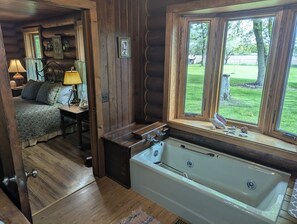 Ein Holzblockhaus-Interieur mit Holzboden, einem Bett mit gemustertem Überwurf, einem kleinen Tisch mit Lampe, einem Fenster mit Blick ins Freie und einer Badewanne.