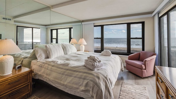 A bedroom with a large bed, a chair, a nightstand, and a view of the ocean through large windows. - Diamond Beach 305 (Ocean City)