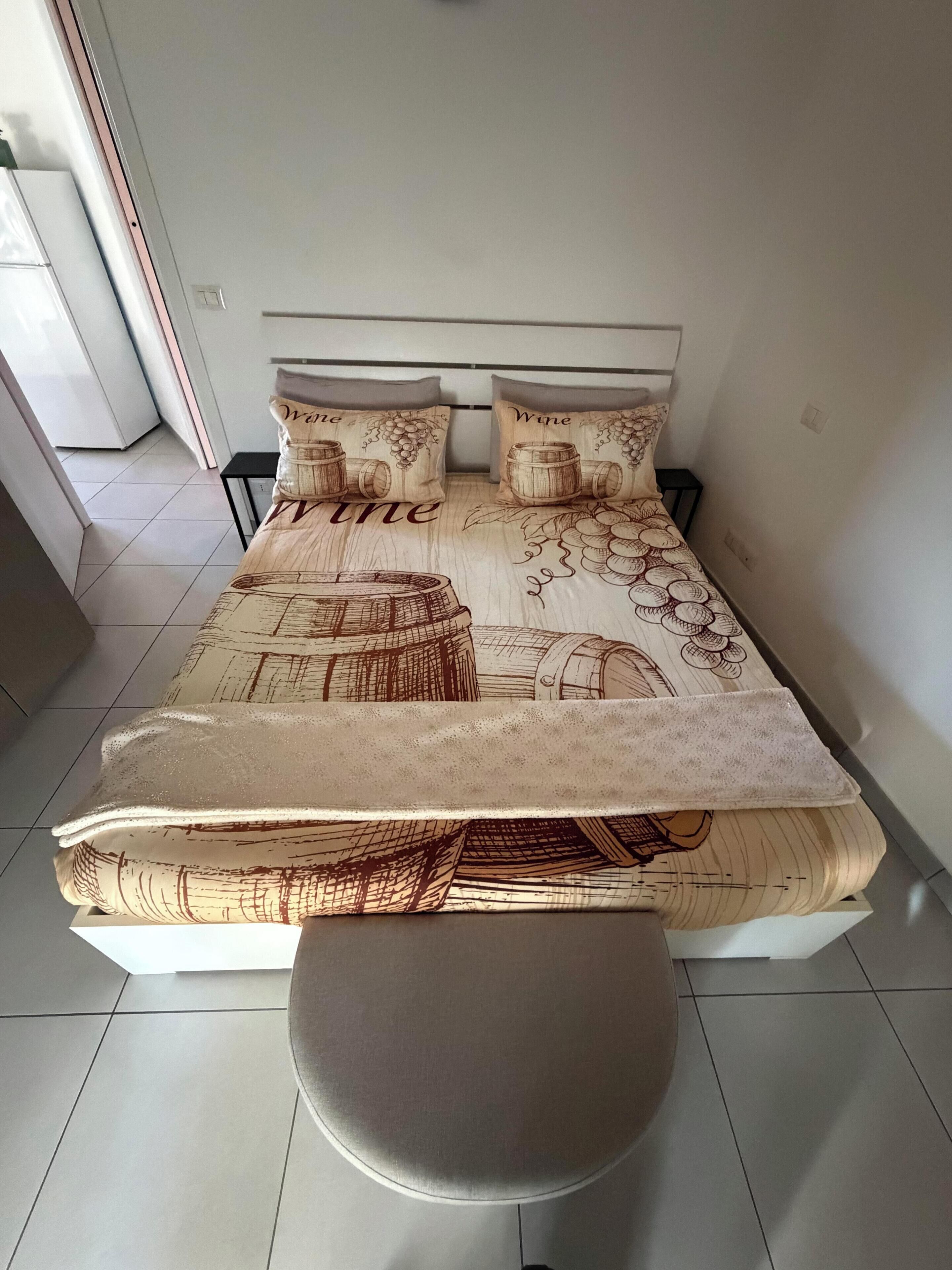 Una cama con una colcha de motivos relacionados al vino, una mesita de noche y una silla.