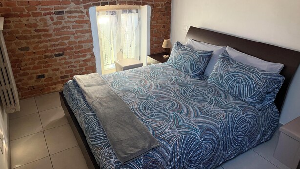 A bedroom with a brick wall, a window with curtains, a bed with patterned bedding, and a bedside table with a lamp. - Casa a Pollenzo (Pollenzo)