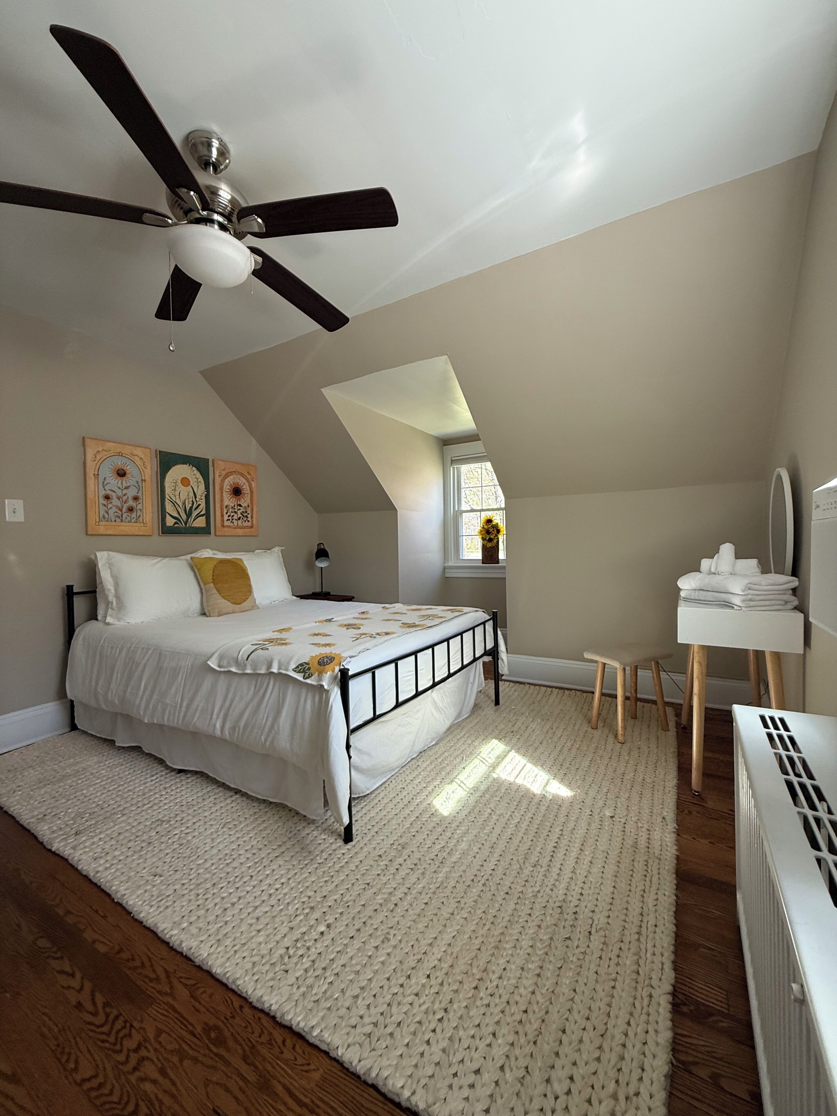 Quarto com cama, ventoinha no teto, radiador, mesa pequena e janela.