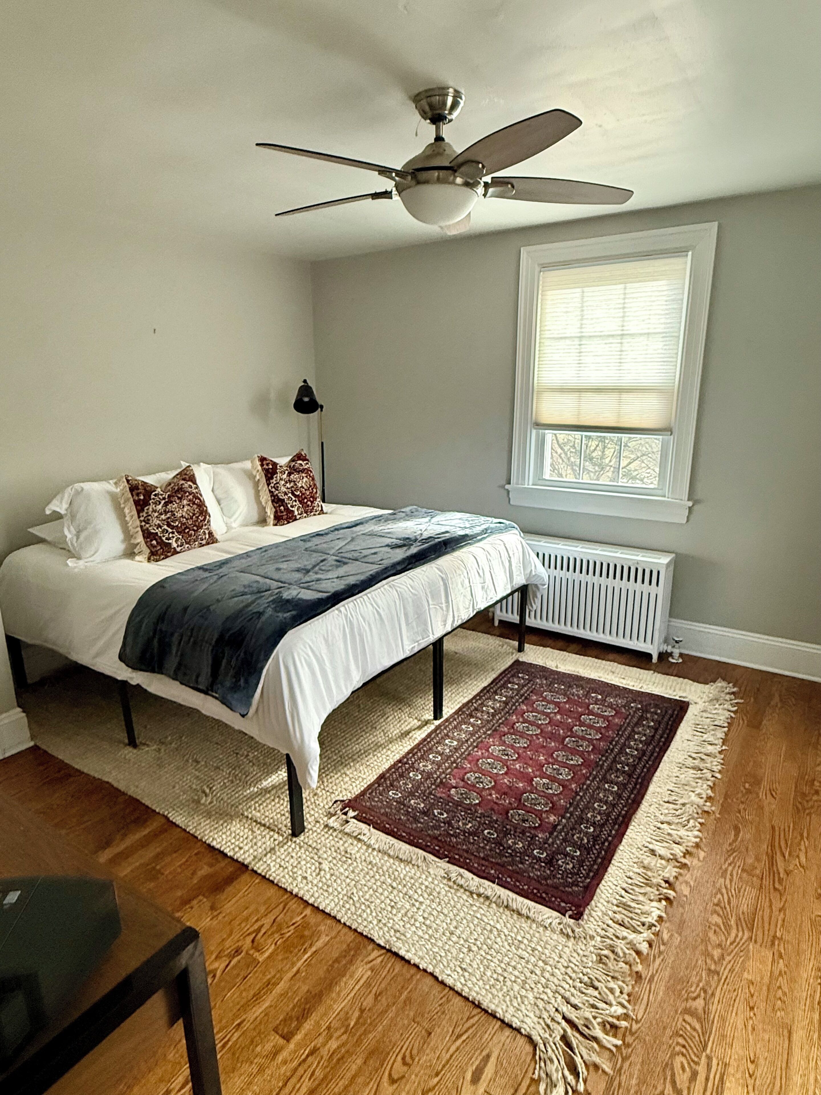 Ein Schlafzimmer mit einem Bett, einem Deckenventilator, einem Fenster mit Jalousien, einem Heizkörper und einem Teppich.