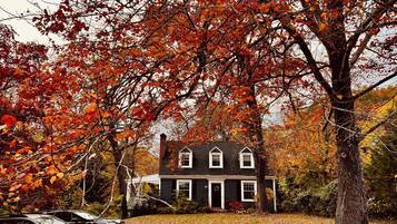 A house with a dark roof, white trim, and a chimney. There are two cars parked in front, and the ground is covered with fallen leaves.