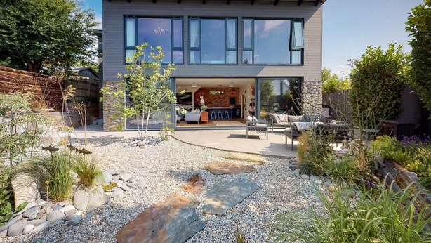 Une maison moderne à deux étages dotée d’un patio comprenant un chemin en pierre, un espace salon extérieur et une variété de plantes.