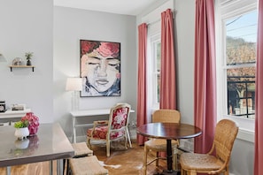 Ein Essbereich mit einem runden Tisch, Korbstühlen, einem rot gepolsterten Sessel, einem Bild an der Wand und einem Fenster mit rosa Vorhängen.