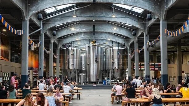 A spacious hall with high ceilings and industrial-style columns, featuring numerous wooden tables and benches, and a central area with large cylindrical structures.