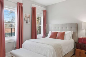 Ein Schlafzimmer mit einem Bett, einem Fenster mit Vorhängen, einem Nachttisch mit einer Lampe und einem Kunstwerk an der Wand.