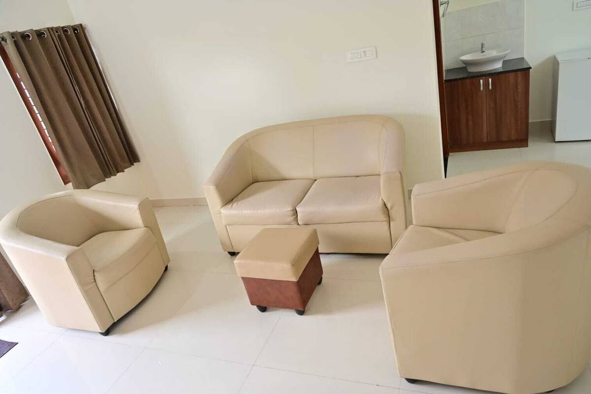 A living room with a beige sofa set, a small ottoman, and a coffee table. There is a window with curtains, a sink, and a cabinet in the background.