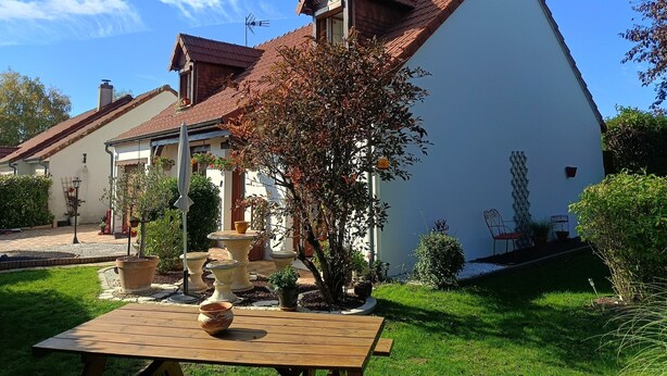 Una casa de dos pisos con techo inclinado, un jardín con plantas en macetas y una mesa de madera en primer plano.