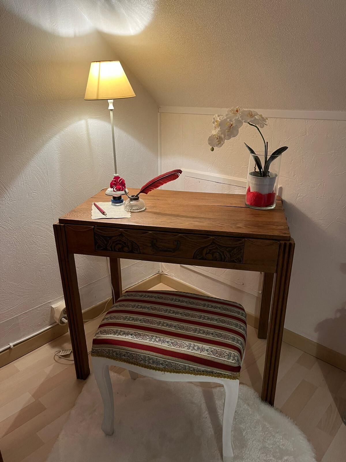Un petit bureau en bois, sur lequel se trouvent une lampe, une plume rouge, un vase en verre contenant une plante et un coussin à motifs.