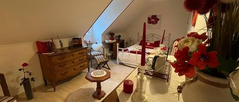 A cozy attic bedroom with a sloped ceiling, a wooden dresser, a chair, a bed with red accents, and a vase with red flowers.