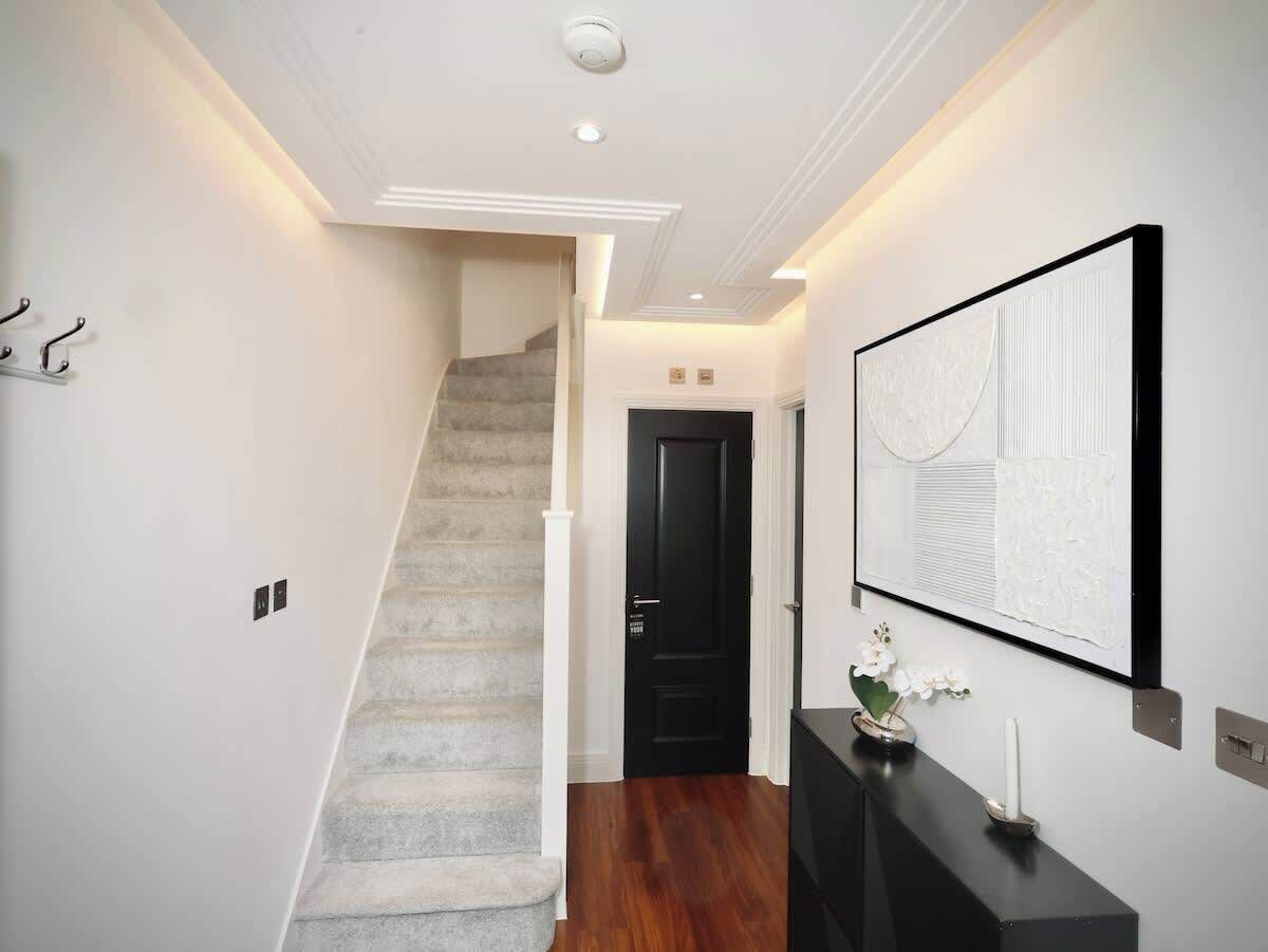 A hallway with a staircase, a black door, a wall-mounted artwork, and a small table with a candle and flowers.