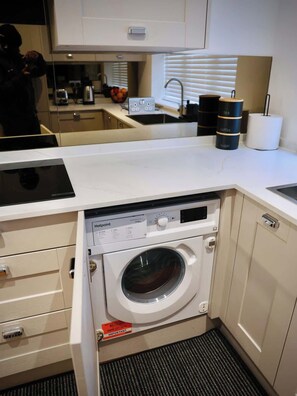Een kleine keuken met een ingebouwde wasmachine en droger, een spoelbak en een aanrecht met een koffieapparaat en theedispenser.