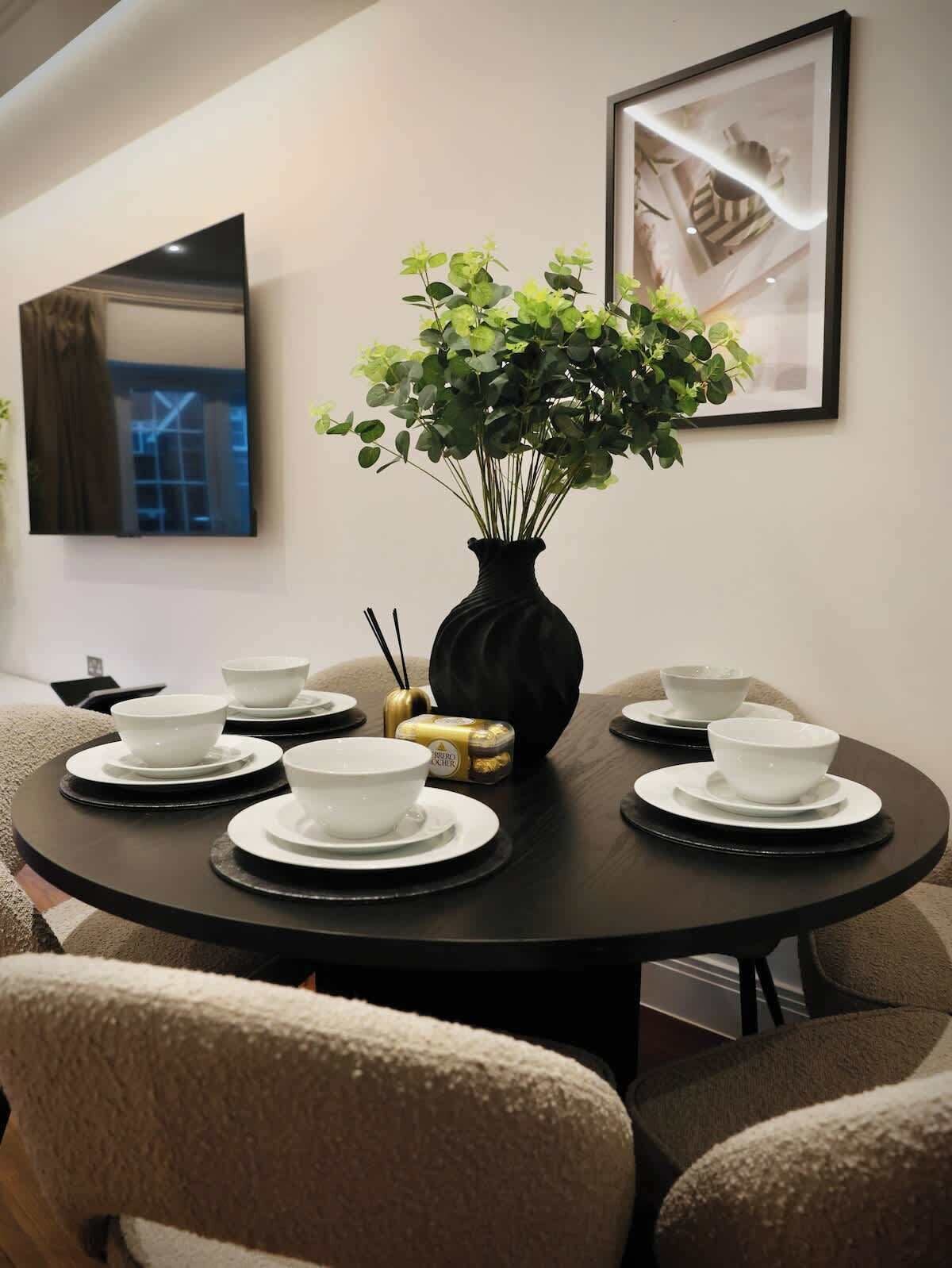 A round dining table with white bowls and black saucers, a black vase with green plants, and a framed picture on the wall.